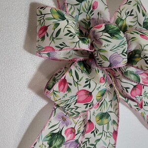 May include: A white ribbon with a pink floral pattern and green leaves. The ribbon is tied into a bow with the ends of the ribbon hanging down.