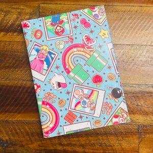 May include: A rectangular notebook with a light blue cover featuring a colorful Super Mario Bros. design. The cover includes images of Mario, Princess Peach, Toad, rainbows, and other iconic elements from the game.