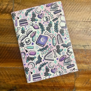 May include: A notebook with a light pink and white cover featuring a repeating pattern of skulls, books, moons, crystals, and potion bottles in purple, teal, and black. The notebook is open, displaying the patterned interior.