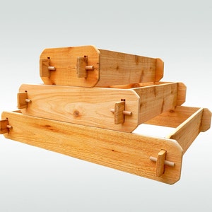 May include: Three wooden raised garden beds stacked on top of each other. The beds are made of natural wood and have a light brown color. The beds are connected with wooden brackets.