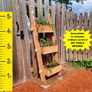 May include: A wooden, three-tiered plant stand with rectangular planters, filled with colorful flowers and greenery. The stand is approximately 5 feet tall and leans against a wooden fence. Text on the image reads: "Assembles in minutes without screws! NO TOOLS NEEDED."