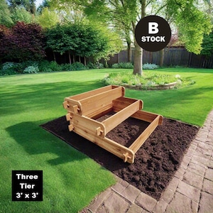 May include: A three-tier wooden raised garden bed in a backyard setting. The bed is made of natural wood and has a rustic look. The bed is 3 feet by 3 feet in size. The text "Three Tier 3' x 3'" is visible in the lower left corner of the image. The text "B STOCK" is visible in the upper right corner of the image.
