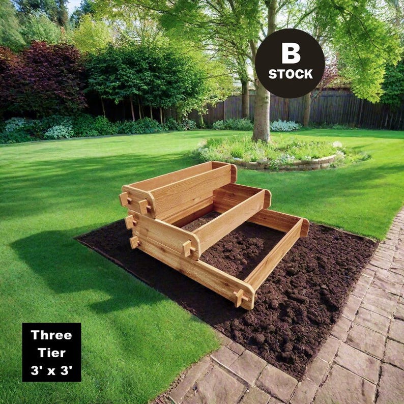May include: A three-tier wooden raised garden bed in a backyard setting. The bed is made of natural wood and has a rustic look. The bed is 3 feet by 3 feet in size. The text "Three Tier 3' x 3'" is visible in the lower left corner of the image. The text "B STOCK" is visible in the upper right corner of the image.