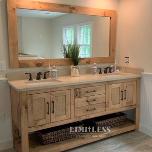May include: A double vanity cabinet made of light-toned wood with a rectangular mirror frame, featuring two sinks, dark metal hardware, three central drawers, two side cabinets, and an open bottom shelf containing two woven baskets for storage.