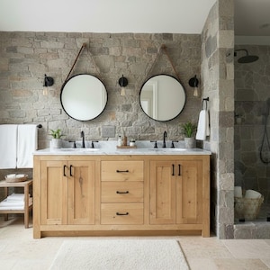 May include: A double vanity bathroom featuring a light wood cabinet with black hardware, a white marble countertop, two round mirrors with leather straps, and a stone wall. A small wooden stool with white towels sits to the left of the vanity.