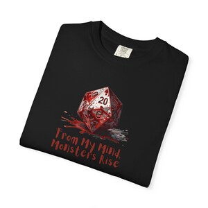 Puede incluir: Camiseta negra con un gráfico de un dado de veinte caras rojo y blanco con salpicaduras de sangre y el texto "From My Mind, Monsters Rise" en rojo. La camiseta está hecha de un material suave.