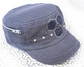 Könnte beinhalten: Eine graue Militärkappe aus Canvas mit einer Reißverschlusstasche an der Seite. Die Kappe hat einen schwarzen Patch mit einem geometrischen Muster und silbernen Nieten am Rand.