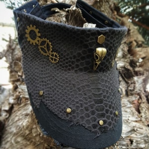 Puede incluir: Una visera negra con un patrón de piel de serpiente falsa y un diseño steampunk. La visera tiene engranajes de metal y un adorno en forma de pájaro.