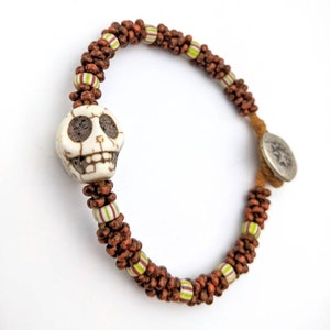May include: A beaded bracelet featuring a white skull charm and brown wooden beads. The bracelet includes green and white striped beads and a silver-colored clasp. The bracelet is designed with a unique, handcrafted aesthetic.