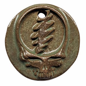 May include: A round, green ceramic pendant with a brown, raised image of the Grateful Dead's iconic Steal Your Face logo. The pendant has a hole at the top for hanging.