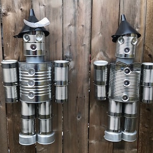 May include: Two tin can robots made from recycled materials. The robots are standing on a wooden fence. The robots have a funnel for a hat and are decorated with silver can lids.