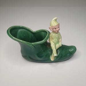May include: A ceramic green shoe-shaped planter with a seated elf figurine. The planter is a deep green color, and the elf is wearing a light green outfit and hat. The elf has a smiling expression.