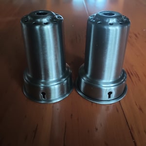 May include: Two stainless steel salt and pepper shakers. Each shaker has a cylindrical body with a flared base and a series of small holes at the top. A keyhole-shaped opening is visible near the base of each shaker.