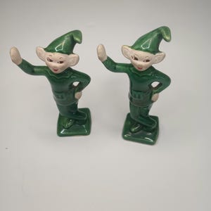 May include: Two ceramic elf figurines. Each elf is wearing a green outfit with a matching hat and a belt. The elves have white skin, large ears, and are smiling. One arm is raised in a wave.