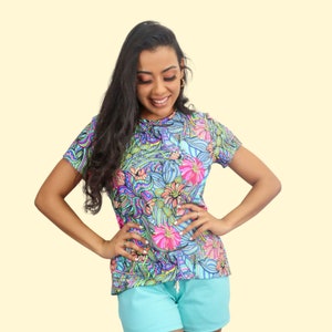May include: A woman wearing a short-sleeved shirt with a colorful floral print. The shirt has a crew neck and is made of a soft, comfortable fabric. The woman is wearing blue shorts and has long, dark hair.