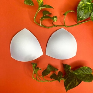 May include: Two white foam bra cups on a bright orange background. The cups are shaped like a triangle with a rounded edge.