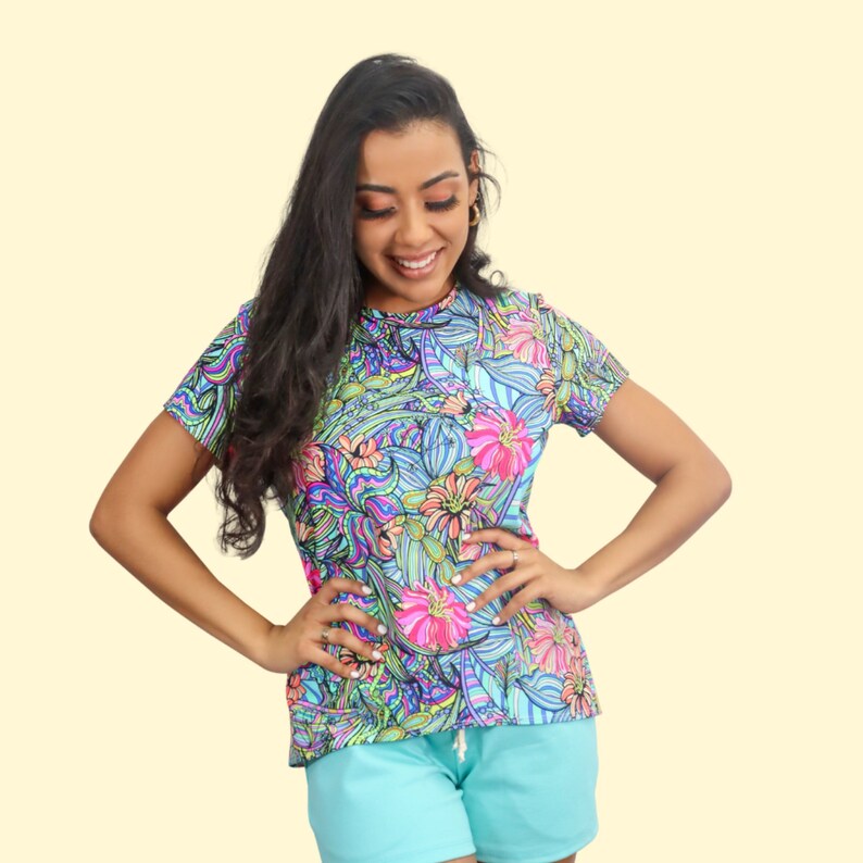 May include: A woman wearing a short-sleeved shirt with a colorful floral print. The shirt has a crew neck and is made of a soft, comfortable fabric. The woman is wearing blue shorts and has long, dark hair.