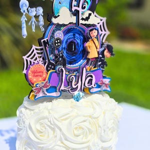May include: A white cake with buttercream frosting, topped with a colorful, layered cake topper. The topper features characters and elements from a fantasy movie, including a girl in a yellow coat, a black cat, and the name "Lyla".