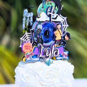 May include: A white cake with a decorative topper featuring a character design. The topper includes the name "Lyla", a spiderweb, a black cat, and a button. The cake is decorated with white frosting roses.
