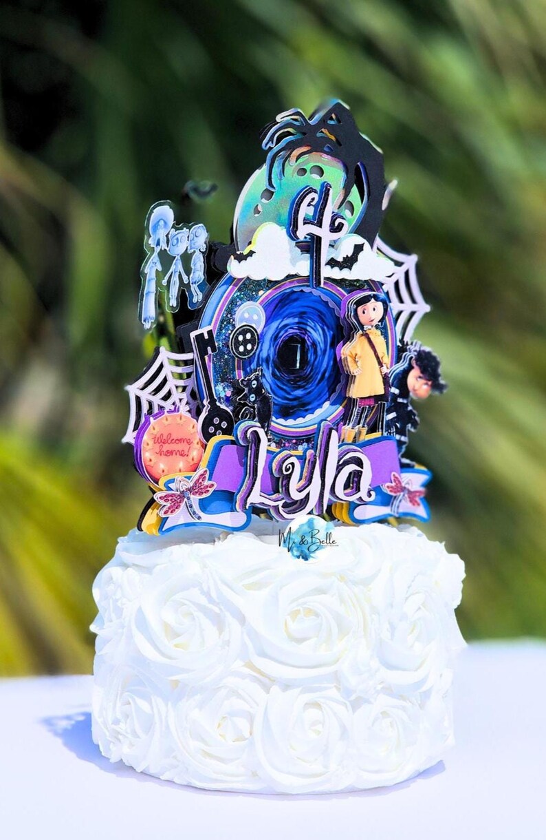 May include: A white cake with a decorative topper featuring a character design. The topper includes the name "Lyla", a spiderweb, a black cat, and a button. The cake is decorated with white frosting roses.
