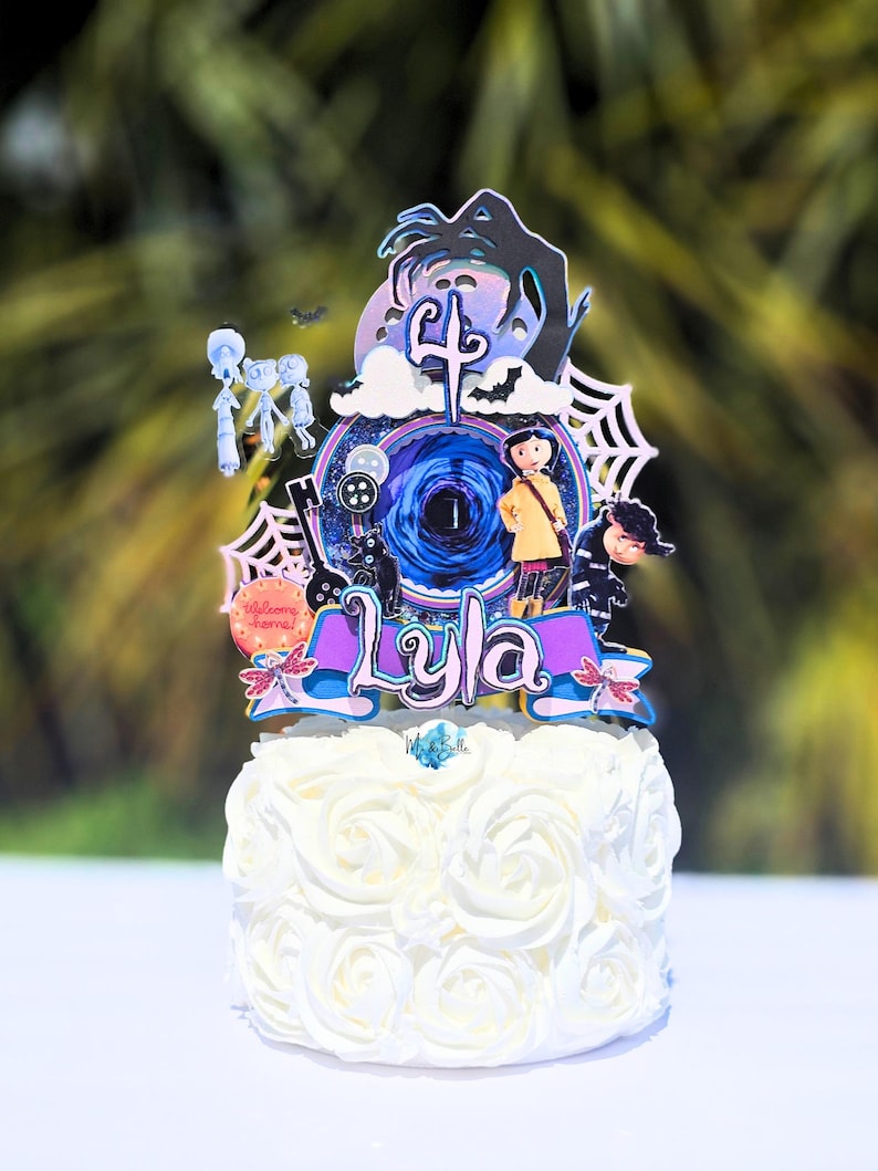 May include: A white cake decorated with white frosting roses, topped with a colorful cake topper. The topper features a character theme with the name "Lyla" and the number "4". The cake is set against a blurred green background.