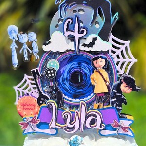 May include: A cake topper featuring characters and elements from the movie "Coraline." The design includes a central portal, the name "Lyla," and various character cutouts, all set against a backdrop of a white cake.