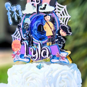 May include: A white cake with a decorative topper featuring characters and elements from the movie "Coraline." The topper includes the name "Lyla," a swirling portal, and other themed details. The cake is decorated with white frosting in a rose pattern.