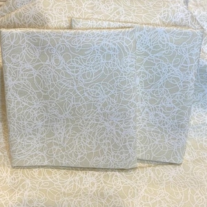 May include: Two beige fabric squares with a white scribble pattern. The squares are stacked on top of each other.