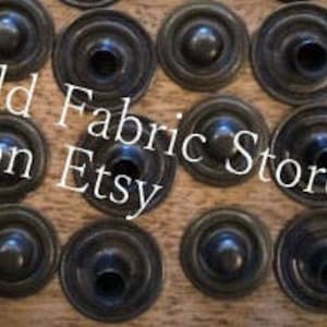 May include: A close-up of 20 black metal snap fasteners. The fasteners are round with a raised center and a hole in the center. The fasteners are arranged in a grid pattern.