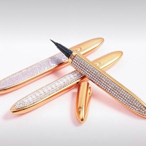 Puede incluir: Tres bolígrafos de delineador de ojos dorados con puntas negras y diamantes de imitación. Los bolígrafos están sobre una superficie rosa.
