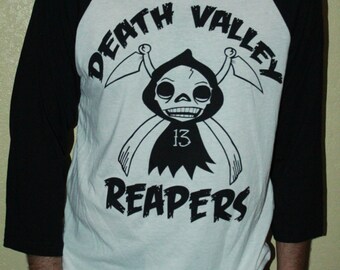 May include: A white and black baseball tee shirt with a graphic of a cartoon grim reaper with the text "Death Valley Reapers" and the number "13" on the front.