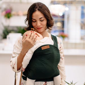 May include: A dark green baby wrap carrier with a brown leather patch that reads "Rick and Bundle". The wrap is being worn by a person holding a baby. The baby is wearing a white long-sleeved shirt.