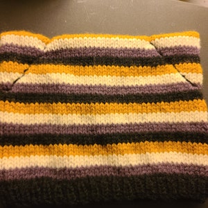May include: A knitted hat with horizontal stripes in shades of black, gold, white, and purple. The hat has a unique shape with small ear-like protrusions at the top. The knitted texture is visible, showcasing the yarn's color variations.
