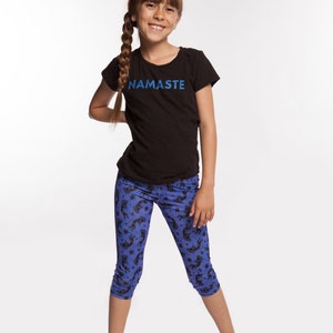 May include: A young person wearing a black t-shirt with the word "NAMASTE" printed on it. They are also wearing blue leggings with a black unicorn pattern.
