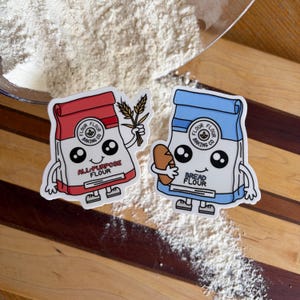 May include: Two die-cut stickers featuring anthropomorphic flour bags. The red bag is labeled ALL-PURPOSE FLOUR and holds wheat stalks. The blue bag is labeled BREAD FLOUR and holds a loaf of bread. Both have large eyes and smiling faces.