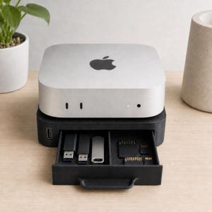 May include: A silver Apple Mac Mini computer sits atop a black storage stand with a pull-out drawer. The drawer contains three USB flash drives and four memory cards of varying sizes. The stand features a single USB port on its left side.