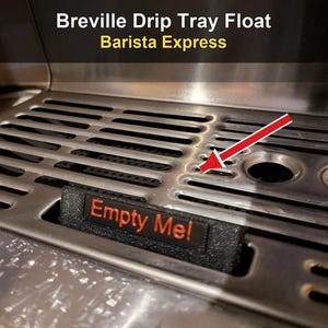 May include: A black plastic drip tray float for a Breville Barista Express espresso machine. The rectangular indicator features the orange text 'Empty Me!' and is positioned within a stainless steel drip tray grate, highlighted by a red arrow.