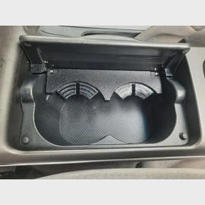 Può includere: Un portabicchieri per auto in plastica nera con due aperture circolari, impugnature interne scanalate e una base antiscivolo con motivo a punti. L'inserto è progettato per essere inserito in una console dotata di coperchio incernierato.