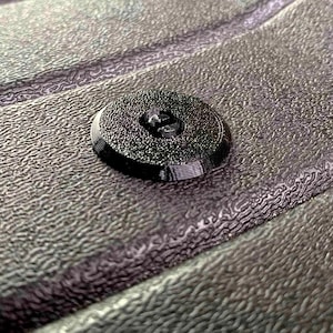 Pode incluir: Um botão preto circular com uma superfície texturizada e dois orifícios centrais. O botão apresenta uma borda ligeiramente elevada e um acabamento fosco, exibindo linhas de camada distintas no perfil lateral, típicas de impressão 3D.