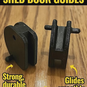 May include: Two black shed door guides are displayed on a wooden surface. The text at the top reads "2x REPLACEMENT SHED DOOR GUIDES". Additional text indicates the guides are made of strong, durable, rust-resistant plastic and glide smoothly.