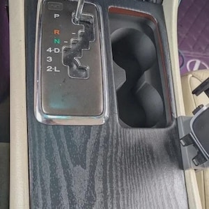 Puede incluir: Una consola central de coche con acabado de veta de madera negra, palanca de cambios con pomo transparente y selector de transmisión automática con marcas P, R, N, 4, 3, 2, L, portavasos doble y un botón etiquetado como PWR ECT SNOW.