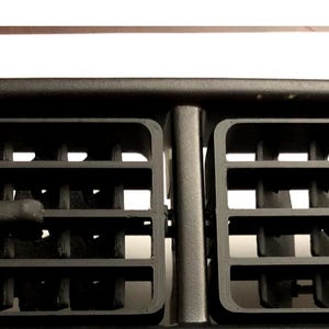 May include: A black automotive dashboard air vent assembly featuring two rectangular sections with horizontal and vertical slats. Each section includes a small, centered adjustment tab for controlling airflow direction, set within a matte black frame.