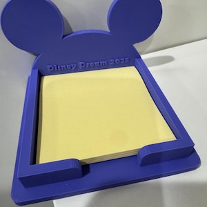 Könnte beinhalten: Ein lila Schreibtisch-Organizer in Form von Mickey Mouse-Ohren, der einen Stapel gelber Haftnotizen enthält. Der Text "Disney Dream 2025" ist auf der Vorderseite sichtbar. Der Organizer besteht aus einem festen Material.