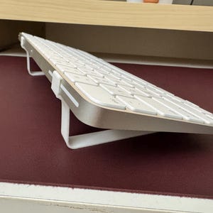 May include: A white wireless keyboard with a silver base is angled upwards on a stand. The stand is made of white plastic and has a simple design.