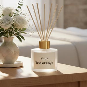 May include: A glass reed diffuser with a gold-colored cap and a label featuring the text Your Text or Logo, accompanied by a ceramic vase containing white roses and baby's breath, all arranged on a wooden surface.