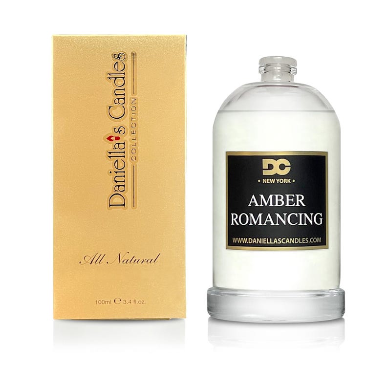 May include: A clear glass diffuser bottle with a black label that reads "AMBER ROMANCING" and "Daniella's Candles Collection." A gold-colored box with the text "All Natural" and "100ml 3.4 fl oz" is next to the bottle.