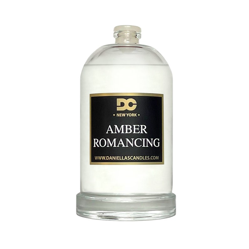 May include: A clear glass bottle with a white liquid inside. The bottle has a black label with gold lettering that reads "AMBER ROMANCING" and "DC NEW YORK". The bottle is sitting on a clear glass base.