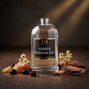 May include: A clear glass candle jar with a black label that reads "AMBER ROMANCING" and "NEW YORK". The jar is surrounded by dried fruit, vanilla beans, and small white flowers. The website www.daniellascandles.com is also visible.