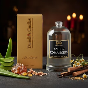 May include: A gold box and a clear glass bottle with a black label that reads "Amber Romancing" are displayed. The box says "Daniella's Candles Collection" and "All Natural". The bottle contains a liquid and is surrounded by botanicals.