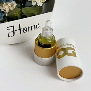 May include: A small glass bottle with a dropper, filled with a yellow liquid, sits next to a white and gold cylindrical container. The container has the text "DG NEW YORK" in gold. The bottle is labeled "1 FL OZ / 30 ML".